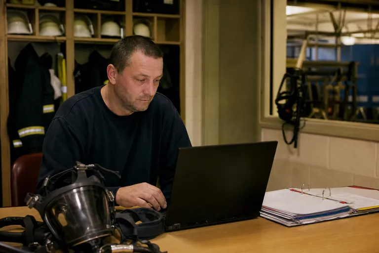 Gerätewart vergleicht am Laptop Fristenübersicht und Aktenordner neben Atemschutzmasken.